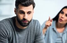 A man with a beard in a grey sweater looks distressed, sitting in the foreground. A woman with long dark hair is in the blurred background, gesturing animatedly as if speaking to him. The atmosphere appears tense.