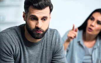 A man with a beard in a grey sweater looks distressed, sitting in the foreground. A woman with long dark hair is in the blurred background, gesturing animatedly as if speaking to him. The atmosphere appears tense.