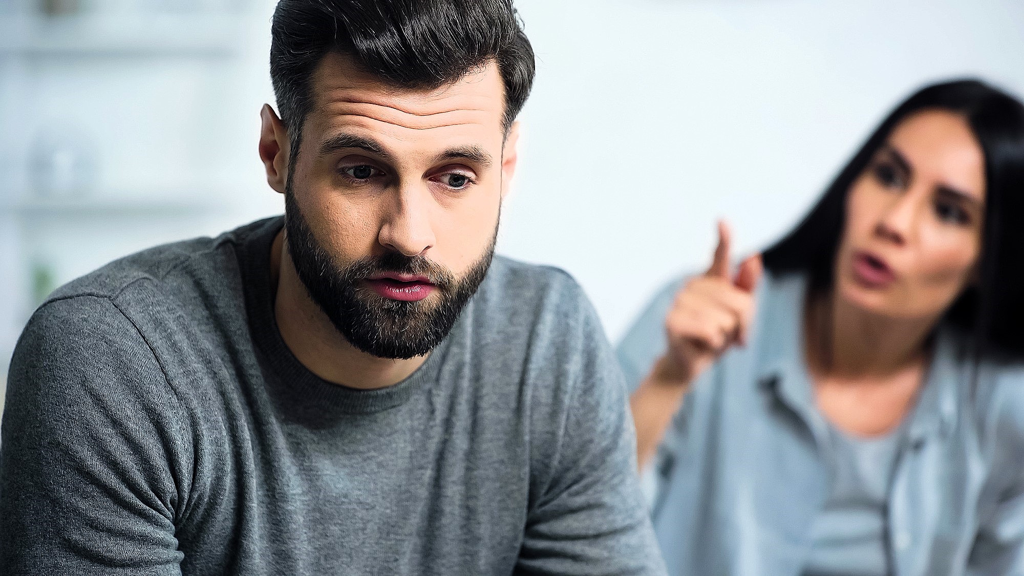 A man with a beard in a grey sweater looks distressed, sitting in the foreground. A woman with long dark hair is in the blurred background, gesturing animatedly as if speaking to him. The atmosphere appears tense.