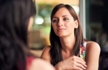 A woman with long brown hair is sitting indoors, wearing a colorful floral top. She is gazing thoughtfully at someone out of frame, with a soft, relaxed expression. The background is softly blurred, suggesting a bright, casual setting.