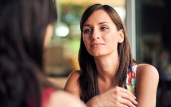 A woman with long brown hair is sitting indoors, wearing a colorful floral top. She is gazing thoughtfully at someone out of frame, with a soft, relaxed expression. The background is softly blurred, suggesting a bright, casual setting.