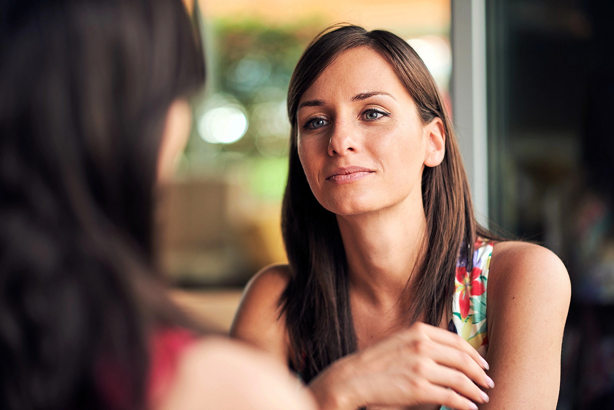 A woman with long brown hair is sitting indoors, wearing a colorful floral top. She is gazing thoughtfully at someone out of frame, with a soft, relaxed expression. The background is softly blurred, suggesting a bright, casual setting.