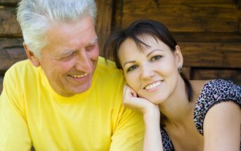 A smiling older man in a yellow shirt sits next to a younger woman with short brown hair, wearing a patterned top. They are outdoors, in front of a wooden background, enjoying a moment together.