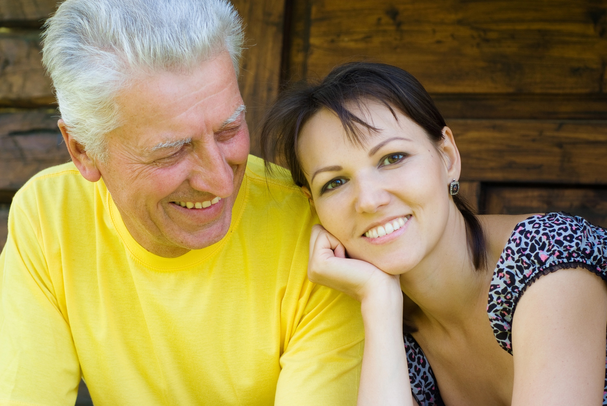 A smiling older man in a yellow shirt sits next to a younger woman with short brown hair, wearing a patterned top. They are outdoors, in front of a wooden background, enjoying a moment together.