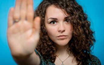 A woman with curly brown hair holds her hand up in a "stop" gesture, her palm facing outward. She has a serious expression, wearing a necklace with a cross pendant. The background is a solid blue color.