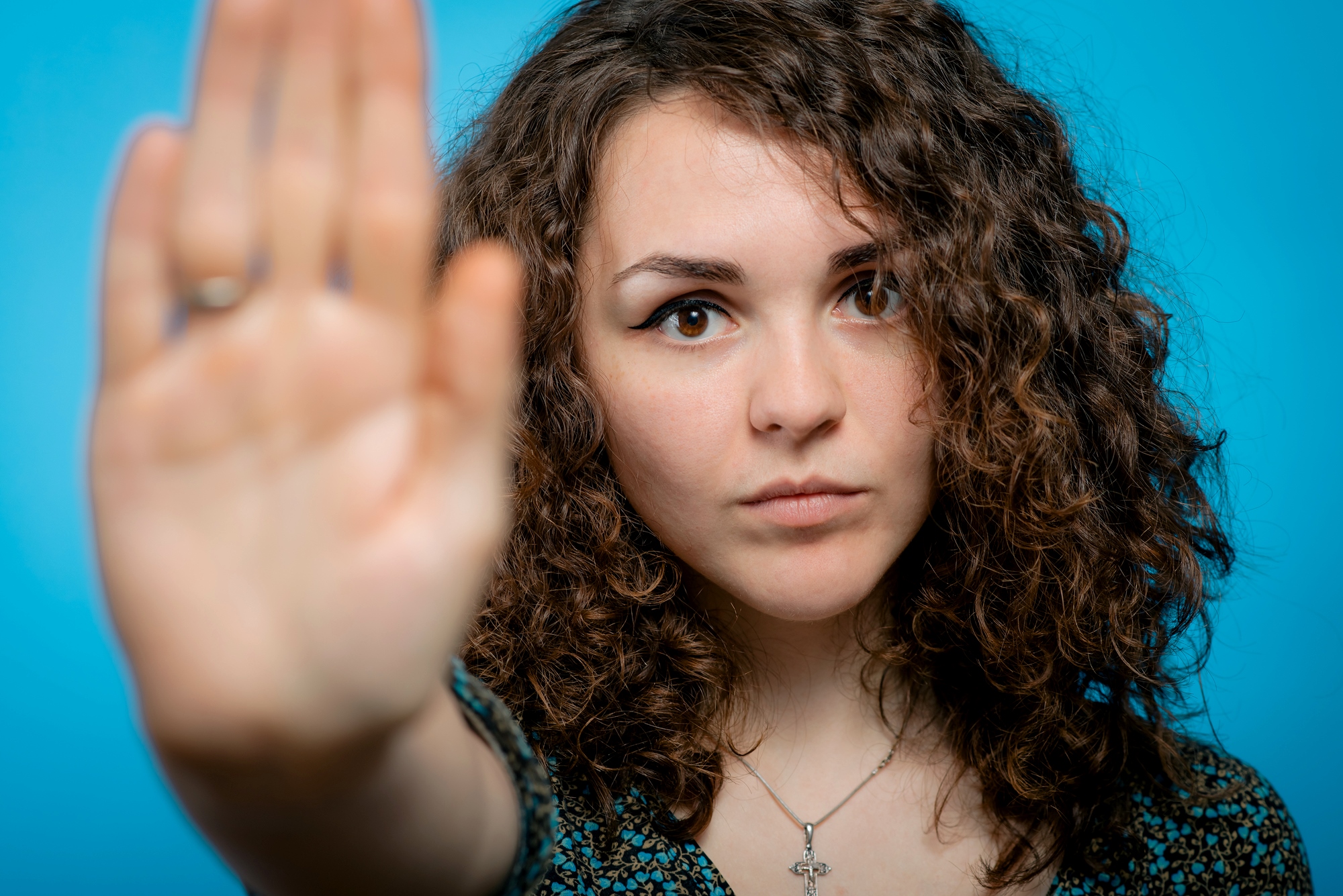 A woman with curly brown hair holds her hand up in a "stop" gesture, her palm facing outward. She has a serious expression, wearing a necklace with a cross pendant. The background is a solid blue color.