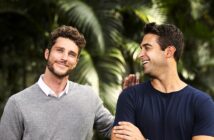 Two men standing outdoors, smiling and having a friendly conversation. One is wearing a gray sweater over a white shirt, and the other is in a blue t-shirt. They are surrounded by lush green foliage, giving a vibrant and relaxed atmosphere.