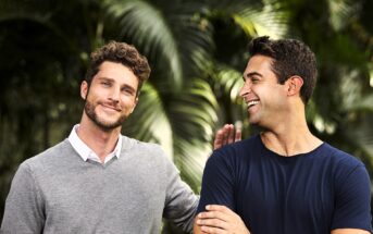 Two men standing outdoors, smiling and having a friendly conversation. One is wearing a gray sweater over a white shirt, and the other is in a blue t-shirt. They are surrounded by lush green foliage, giving a vibrant and relaxed atmosphere.