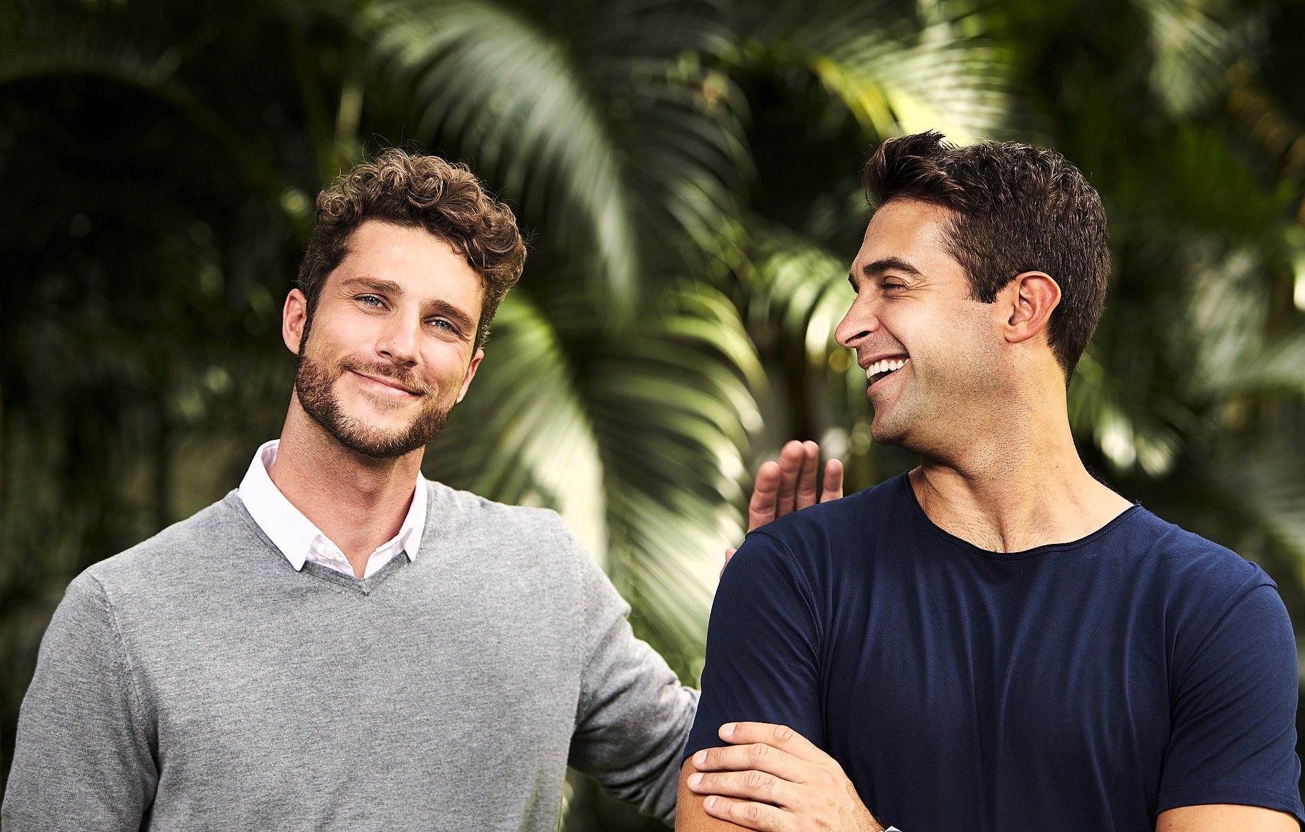 Two men standing outdoors, smiling and having a friendly conversation. One is wearing a gray sweater over a white shirt, and the other is in a blue t-shirt. They are surrounded by lush green foliage, giving a vibrant and relaxed atmosphere.