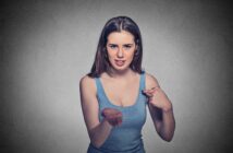 A woman with long brown hair, wearing a blue tank top, stands against a gray background. She points to herself with one hand while her other hand is extended forward, palm up, with a confused or questioning expression on her face.