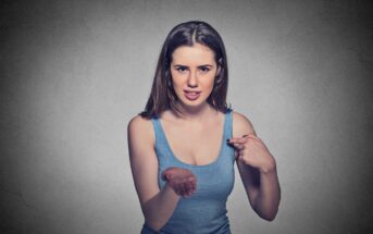 A woman with long brown hair, wearing a blue tank top, stands against a gray background. She points to herself with one hand while her other hand is extended forward, palm up, with a confused or questioning expression on her face.