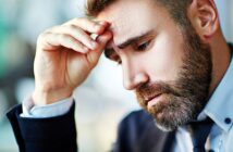 A man with a beard looks thoughtful, resting his head on his hand. He is wearing a suit and appears deep in concentration or contemplation. The background is blurred, suggesting an indoor setting.