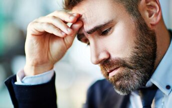 A man with a beard looks thoughtful, resting his head on his hand. He is wearing a suit and appears deep in concentration or contemplation. The background is blurred, suggesting an indoor setting.