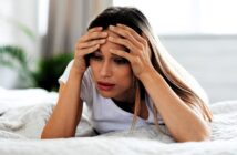 A woman with long hair, wearing a white t-shirt, lies on a bed with a concerned expression. She rests her head in her hands, looking downward. The background is softly blurred, suggesting a serene indoor setting with some greenery.