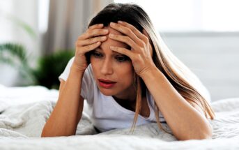 A woman with long hair, wearing a white t-shirt, lies on a bed with a concerned expression. She rests her head in her hands, looking downward. The background is softly blurred, suggesting a serene indoor setting with some greenery.