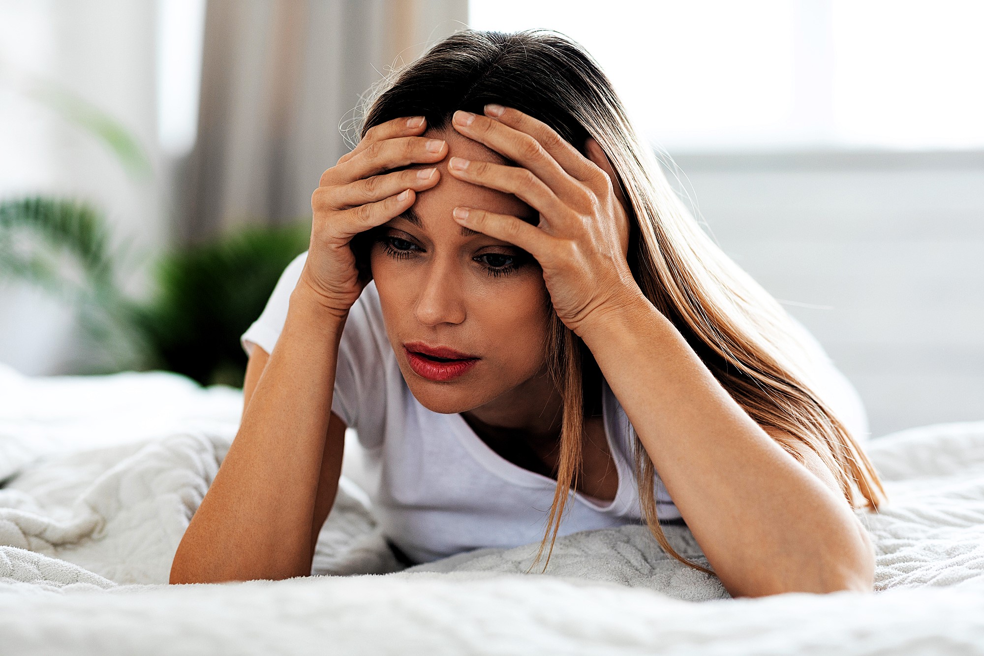 A woman with long hair, wearing a white t-shirt, lies on a bed with a concerned expression. She rests her head in her hands, looking downward. The background is softly blurred, suggesting a serene indoor setting with some greenery.