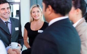 A group of business professionals are engaged in a conversation. The woman in the center is smiling, and a man is holding a cup. They are wearing name tags, and the setting appears to be a networking event or conference.