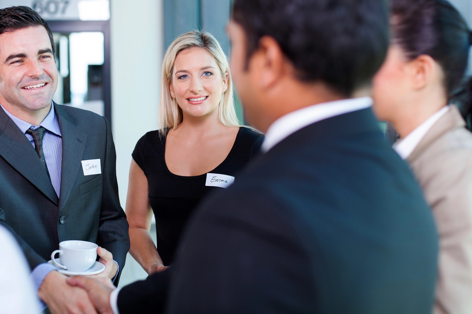 A group of business professionals are engaged in a conversation. The woman in the center is smiling, and a man is holding a cup. They are wearing name tags, and the setting appears to be a networking event or conference.