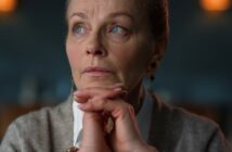 A woman with light brown hair tied back is gazing thoughtfully to the side. She rests her chin on her clasped hands. She wears a white shirt and gray sweater, with earrings visible. The background is softly blurred.