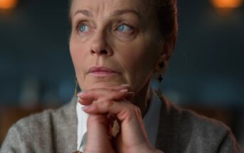 A woman with light brown hair tied back is gazing thoughtfully to the side. She rests her chin on her clasped hands. She wears a white shirt and gray sweater, with earrings visible. The background is softly blurred.