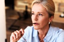 A thoughtful woman with blonde hair holds the arm of her glasses near her lips, looking contemplative. She wears a light blue shirt and pearl earrings, seated in an indoor setting with blurred wooden decor in the background.