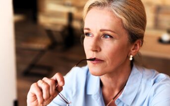 A thoughtful woman with blonde hair holds the arm of her glasses near her lips, looking contemplative. She wears a light blue shirt and pearl earrings, seated in an indoor setting with blurred wooden decor in the background.