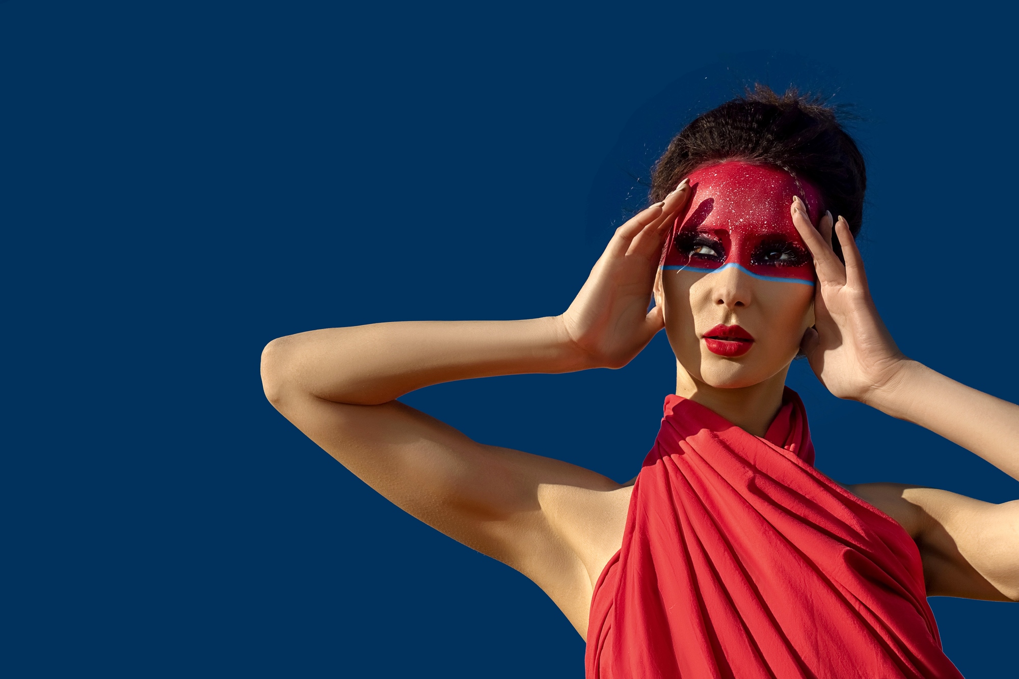 A woman poses against a solid blue background, wearing a red dress. Her face is adorned with red face paint around her eyes and forehead, and she holds her hands near her head in a striking pose.