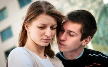 A young woman with long, brown hair looks down with a concerned expression while a young man leans close to her, appearing to speak softly. They are standing outdoors with buildings in the background.