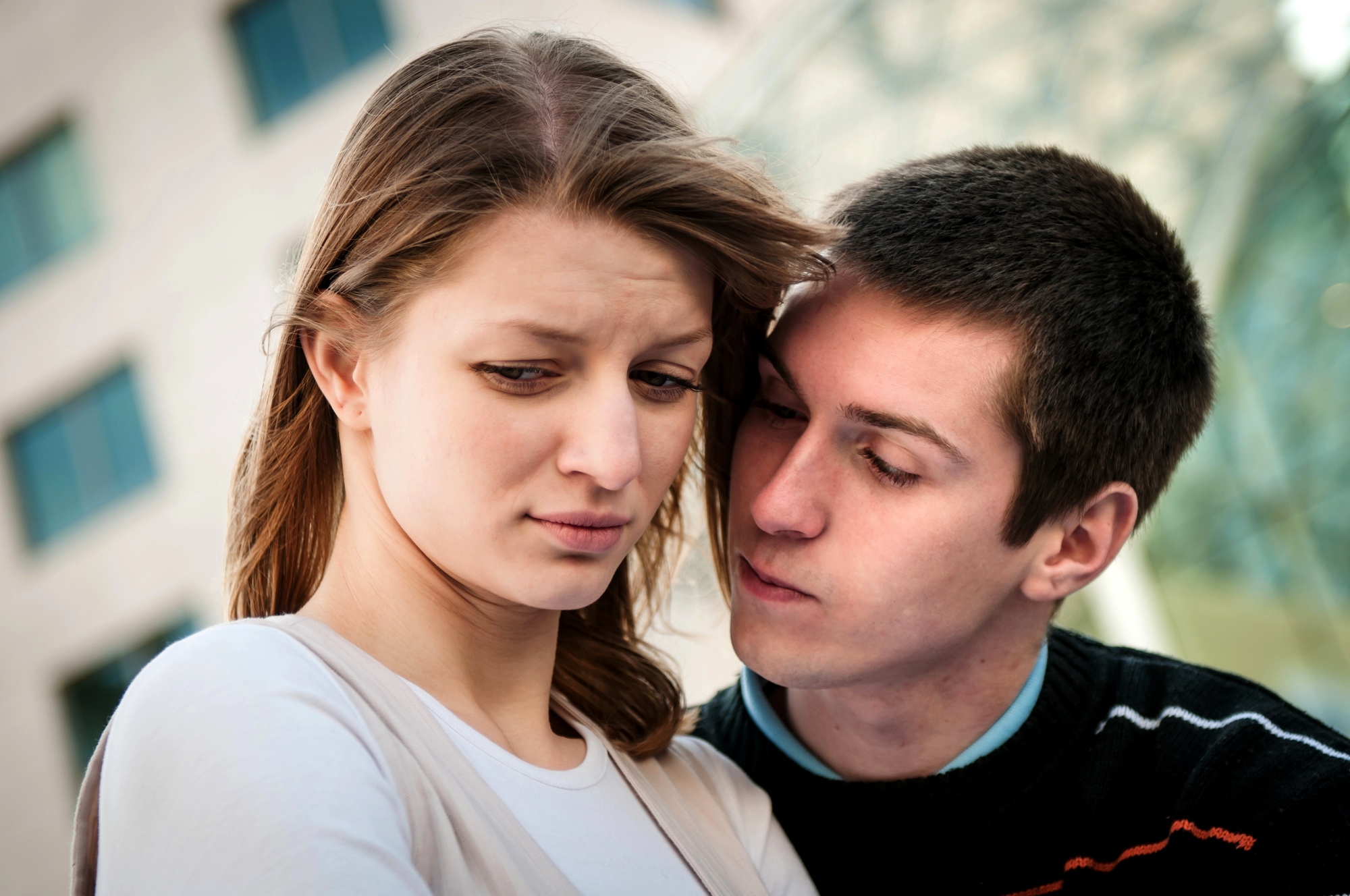 A young woman with long, brown hair looks down with a concerned expression while a young man leans close to her, appearing to speak softly. They are standing outdoors with buildings in the background.