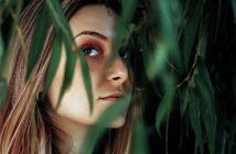 A woman partially concealed by lush green leaves looks over her shoulder. Her face is softly lit, highlighting her eyes and natural makeup. The greenery adds a sense of mystery and nature to the scene.