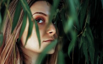 A woman partially concealed by lush green leaves looks over her shoulder. Her face is softly lit, highlighting her eyes and natural makeup. The greenery adds a sense of mystery and nature to the scene.