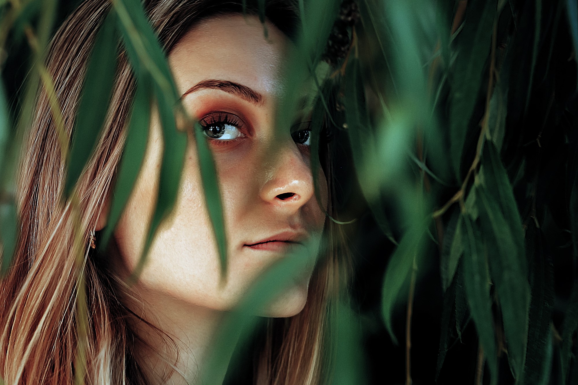 A woman partially concealed by lush green leaves looks over her shoulder. Her face is softly lit, highlighting her eyes and natural makeup. The greenery adds a sense of mystery and nature to the scene.