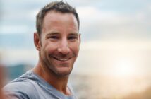 Smiling man in a gray shirt standing outdoors with a blurred cityscape and sunset in the background.