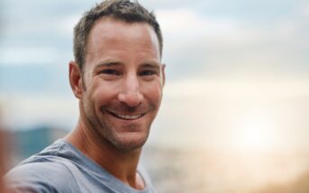 Smiling man in a gray shirt standing outdoors with a blurred cityscape and sunset in the background.