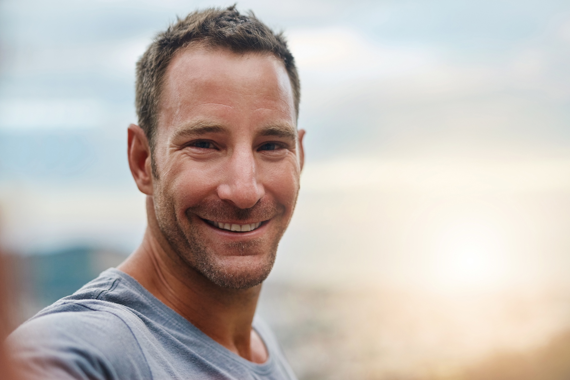 Smiling man in a gray shirt standing outdoors with a blurred cityscape and sunset in the background.