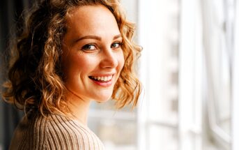 A person with curly hair is smiling and looking over their shoulder by a window. They're wearing a light-colored sweater, and natural light is illuminating their face. The background is softly blurred.