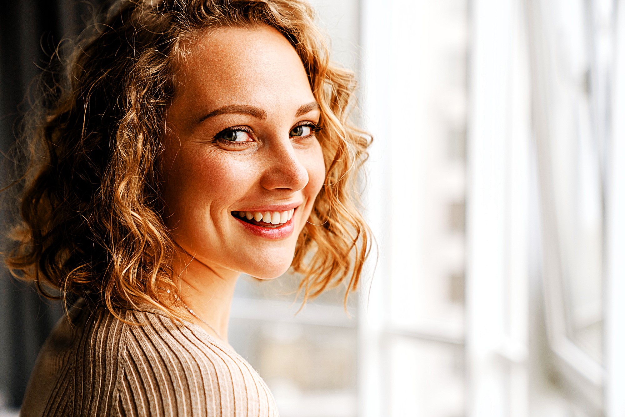 A person with curly hair is smiling and looking over their shoulder by a window. They're wearing a light-colored sweater, and natural light is illuminating their face. The background is softly blurred.