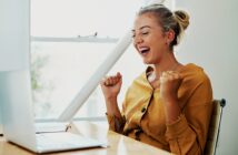 A woman with blonde hair in a bun is sitting at a desk, looking at a laptop with excitement. She is wearing a mustard-colored shirt and has her fists raised in celebration. The setting is bright with natural light coming through a window.