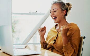 A woman with blonde hair in a bun is sitting at a desk, looking at a laptop with excitement. She is wearing a mustard-colored shirt and has her fists raised in celebration. The setting is bright with natural light coming through a window.