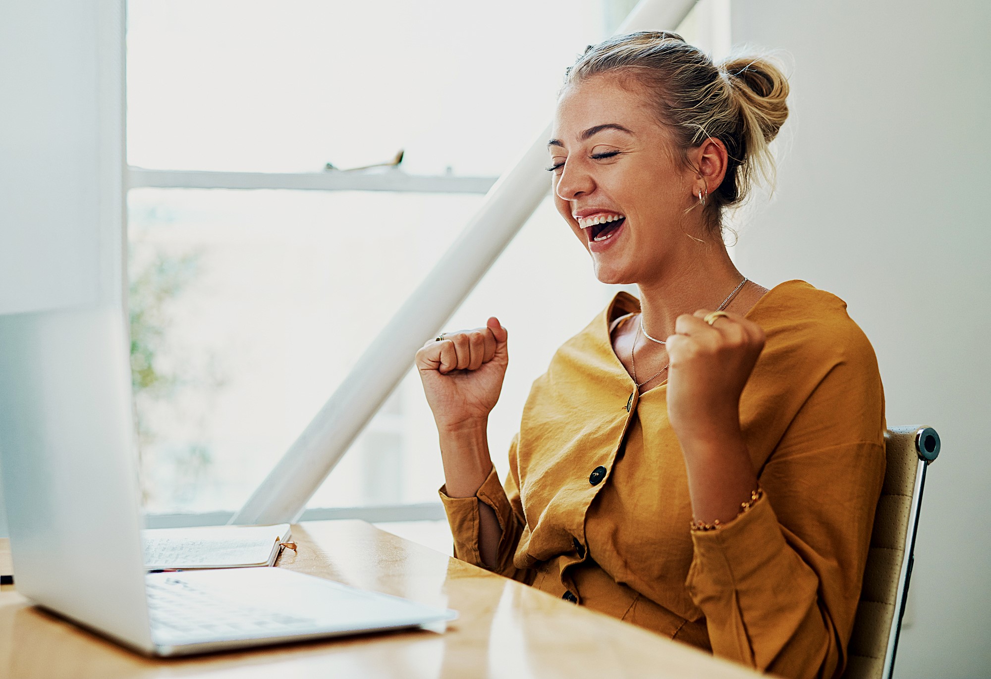 A woman with blonde hair in a bun is sitting at a desk, looking at a laptop with excitement. She is wearing a mustard-colored shirt and has her fists raised in celebration. The setting is bright with natural light coming through a window.