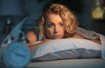 A woman with curly blonde hair lies awake on her bed, resting her chin on a pillow, appearing thoughtful. A blurred alarm clock is visible in the foreground, suggesting early morning or late night.