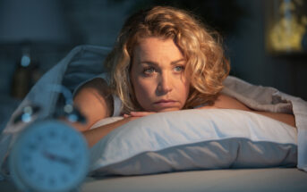 A woman with curly blonde hair lies awake on her bed, resting her chin on a pillow, appearing thoughtful. A blurred alarm clock is visible in the foreground, suggesting early morning or late night.