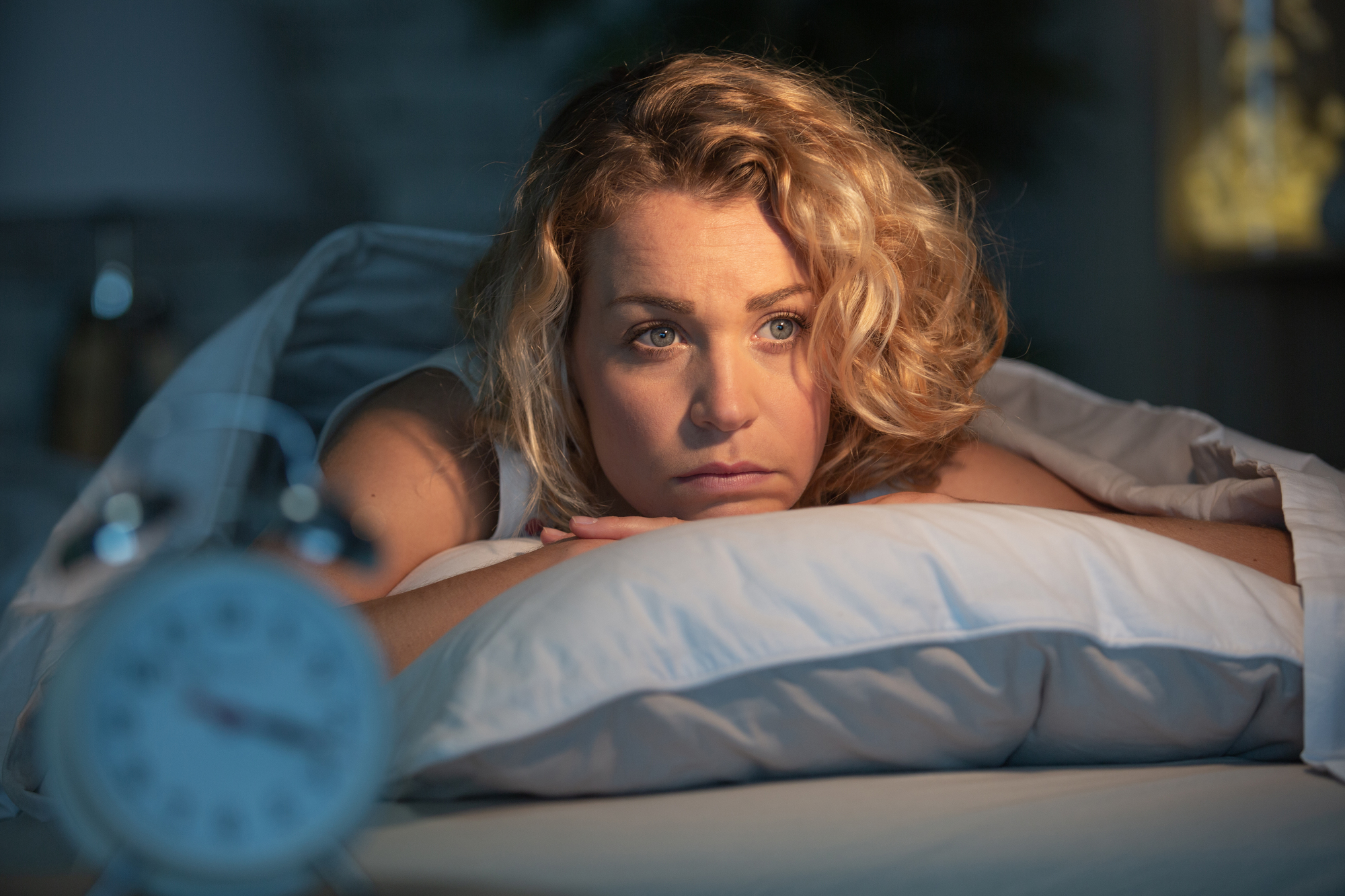 A woman with curly blonde hair lies awake on her bed, resting her chin on a pillow, appearing thoughtful. A blurred alarm clock is visible in the foreground, suggesting early morning or late night.