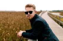 A person with sunglasses and a dark jacket leans on a wooden railing along a boardwalk. They are surrounded by tall grasses, and the atmosphere is calm and overcast.