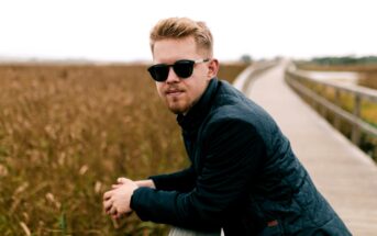 A person with sunglasses and a dark jacket leans on a wooden railing along a boardwalk. They are surrounded by tall grasses, and the atmosphere is calm and overcast.