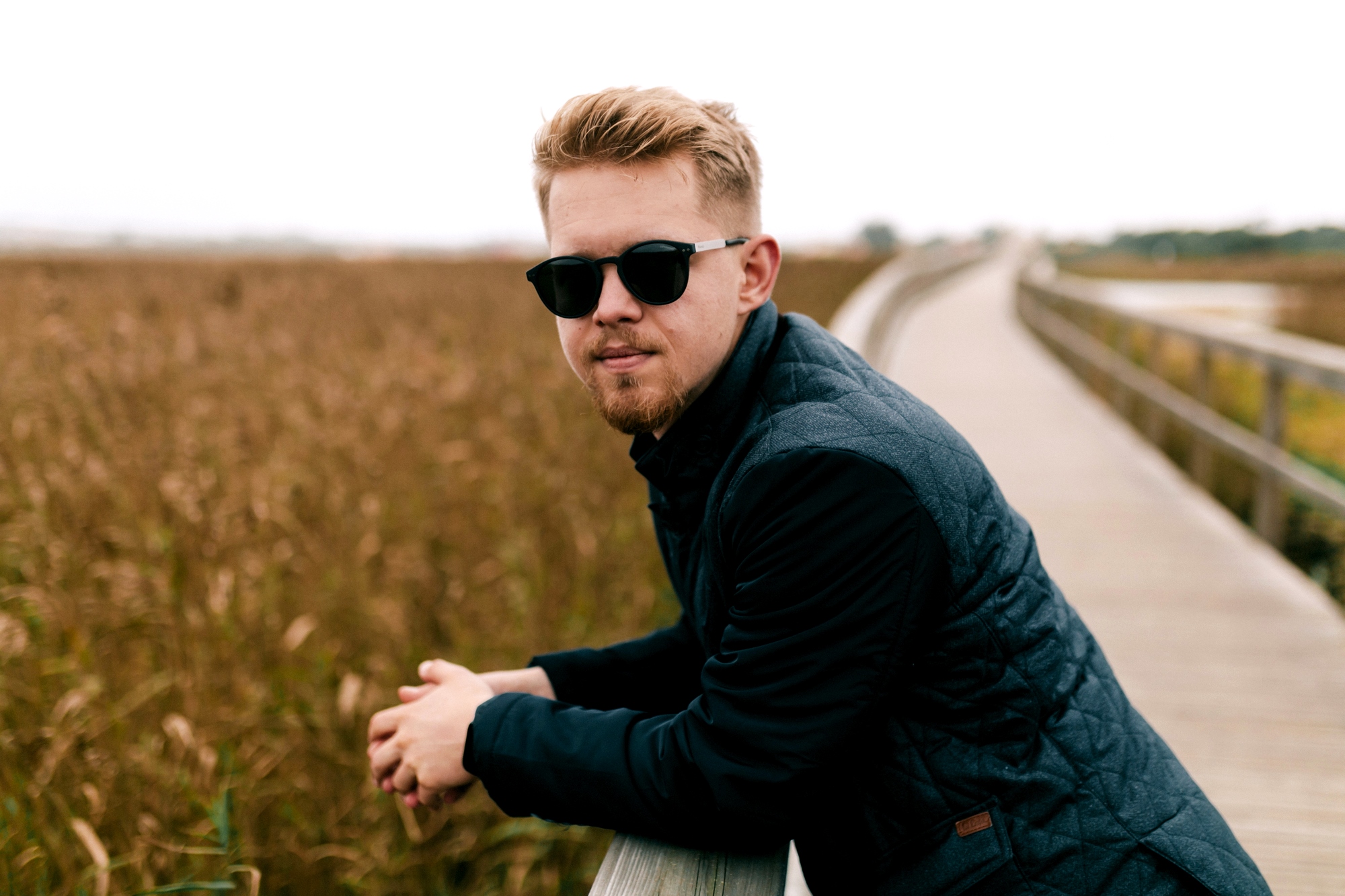 A person with sunglasses and a dark jacket leans on a wooden railing along a boardwalk. They are surrounded by tall grasses, and the atmosphere is calm and overcast.