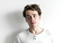 A person with wavy hair wearing a white henley shirt leans against a plain white wall, looking to the side with a contemplative expression.