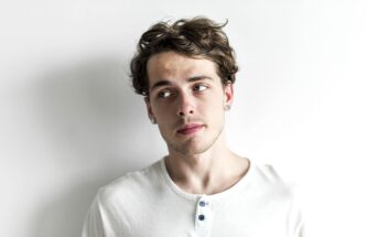 A person with wavy hair wearing a white henley shirt leans against a plain white wall, looking to the side with a contemplative expression.