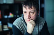 A man with dark hair and facial hair, wearing a striped sweater, is seated indoors. He rests his chin on his hand, appearing thoughtful, with a blurred background featuring shelves and books.