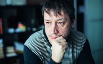 A man with dark hair and facial hair, wearing a striped sweater, is seated indoors. He rests his chin on his hand, appearing thoughtful, with a blurred background featuring shelves and books.